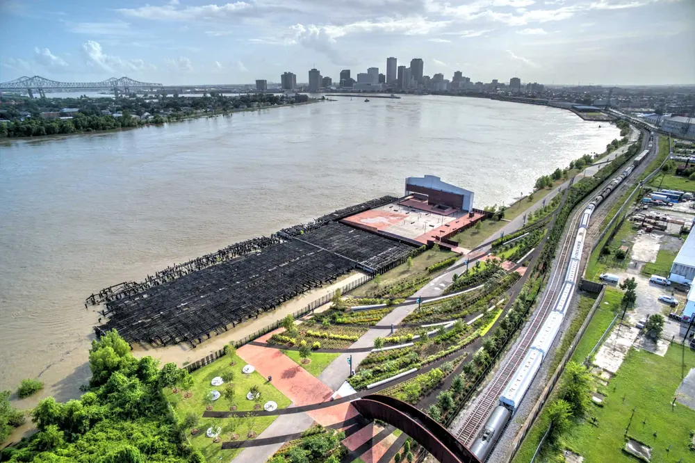 Aerial view of Crescent Park: a landscaped riverfront and old wharf along the Mississippi, with the downtown skyline in the distance.