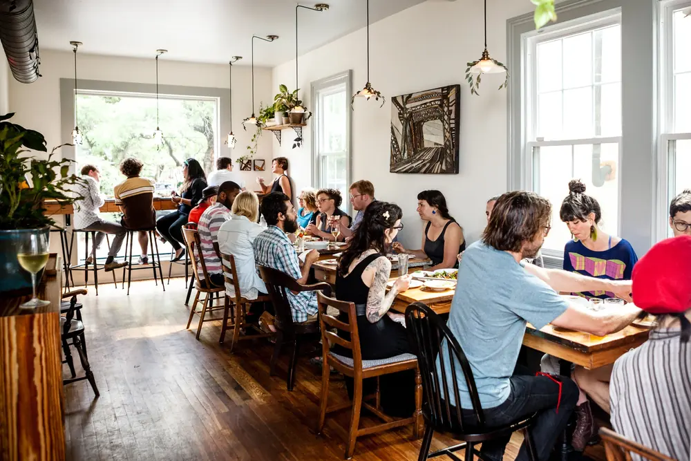 Diners at long wooden tables in the bright dining room of 1000 Figs, with tall windows and live oaks outside.