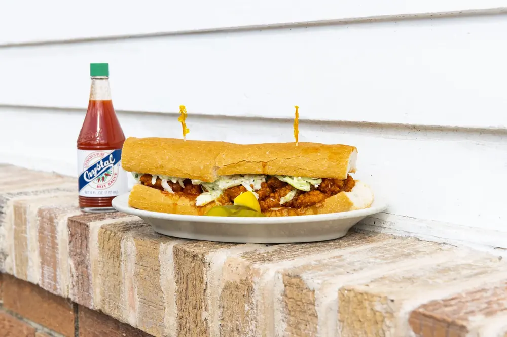 A BBQ shrimp po-boy on a plate on a brick ledge beside a bottle of Crystal hot sauce.