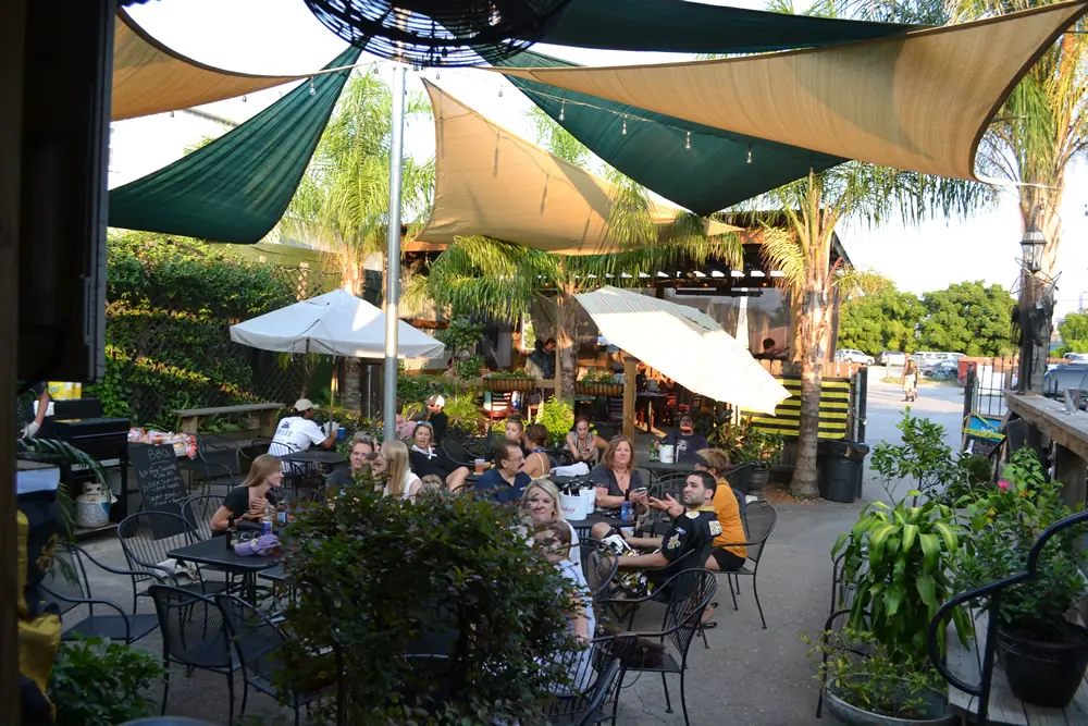 People at tables in the shaded backyard patio of Bayou Beer Garden, under sail-cloth shades and palms.