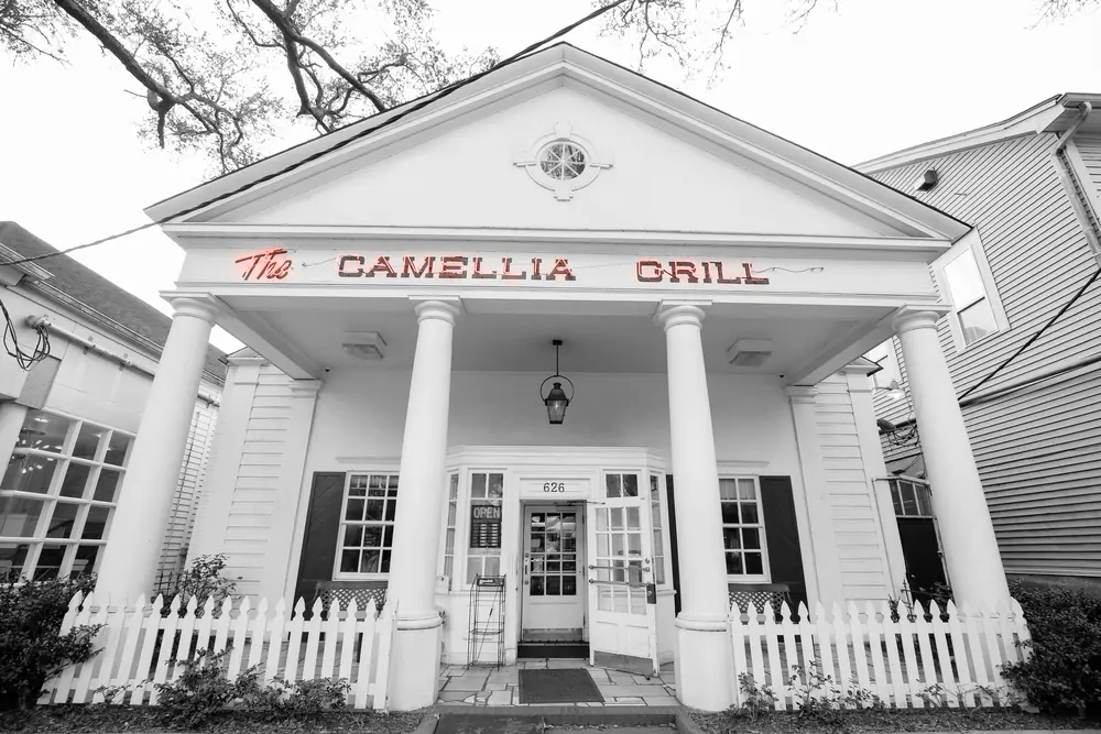 A black-and-white photo of the white-columned front of Camellia Grill, with red lettering and a white picket fence.