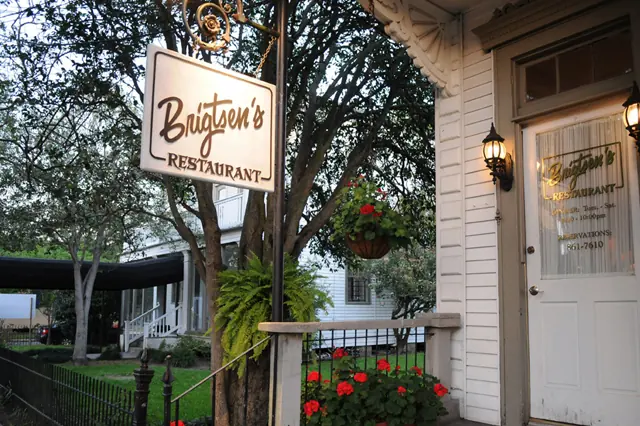 The sign and front door of Brigtsen’s, a white wooden cottage with hanging baskets and red geraniums.