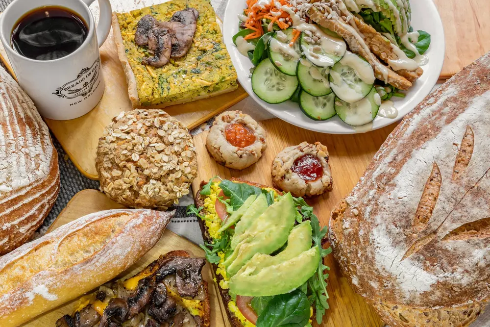 A wooden board of breads, pastries, an avocado and mushroom sandwich, a salad and a cup of coffee.