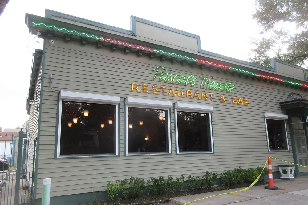 The green clapboard side of Pascal’s Manale with neon script reading Pascal’s Manale Restaurant and Bar.