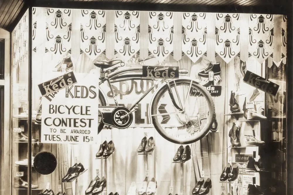 A black-and-white photo of a Haase’s shop window with a Keds bicycle contest sign, a bicycle and rows of shoes.